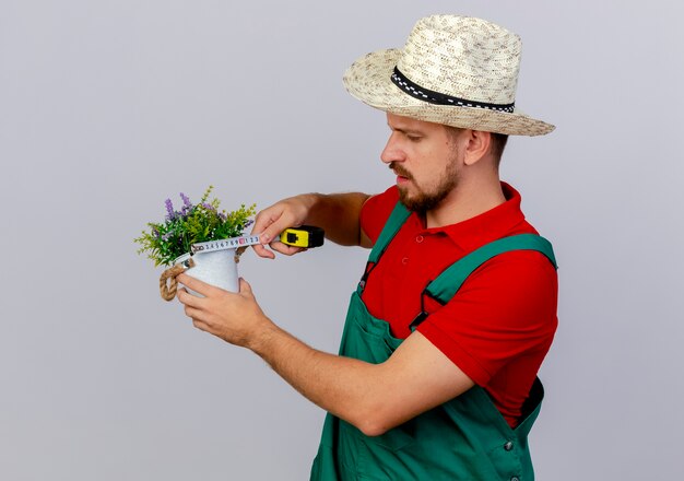 Joven jardinero eslavo guapo concentrado en uniforme y sombrero sosteniendo maceta y medidor de cinta midiéndolo aislado