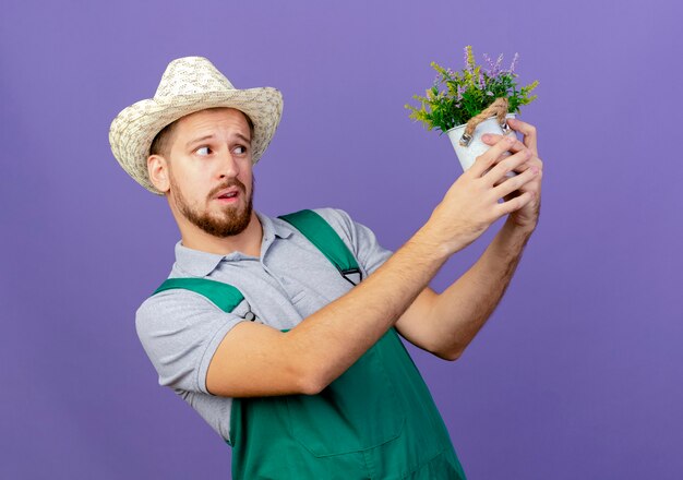 Joven jardinero eslavo guapo dudoso en uniforme y sombrero sosteniendo y mirando maceta aislada en la pared púrpura