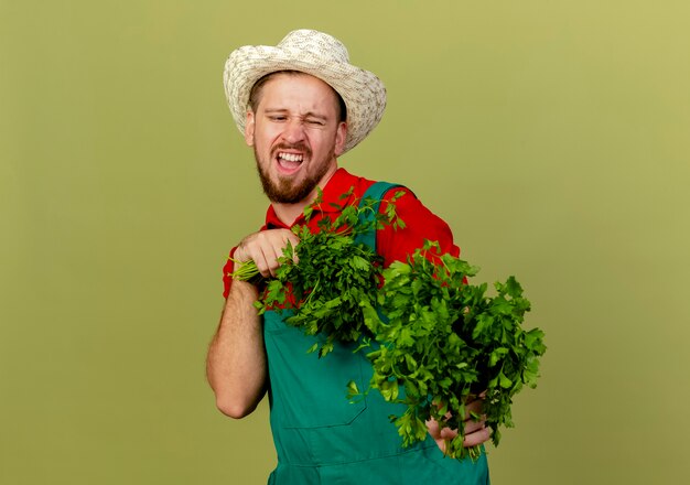 Joven jardinero eslavo guapo molesto en uniforme y sombrero sosteniendo y estirando cilantro mirándolo aislado en la pared verde oliva con espacio de copia