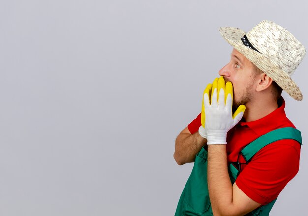 Joven jardinero eslavo guapo sorprendido en uniforme y sombrero con guantes de jardinero de pie en la vista de perfil manteniendo las manos en la boca mirando directamente aislado