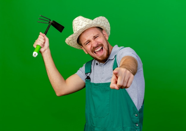 Joven jardinero eslavo guapo en uniforme y sombrero mirando y apuntando sosteniendo azada y gritando