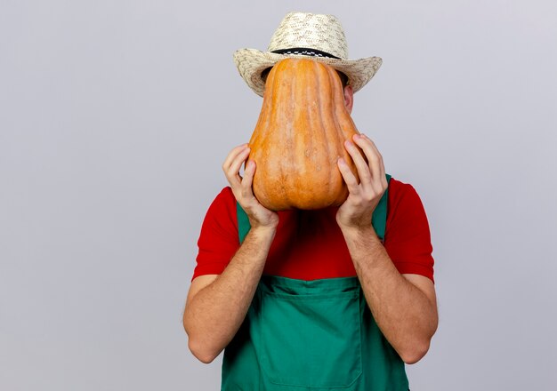 Joven jardinero eslavo guapo en uniforme y sombrero sosteniendo y escondido detrás de calabaza aislado