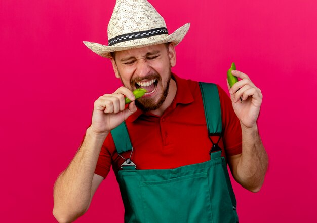 Joven jardinero eslavo guapo en uniforme y sombrero sosteniendo y mordiendo pimienta con los ojos cerrados