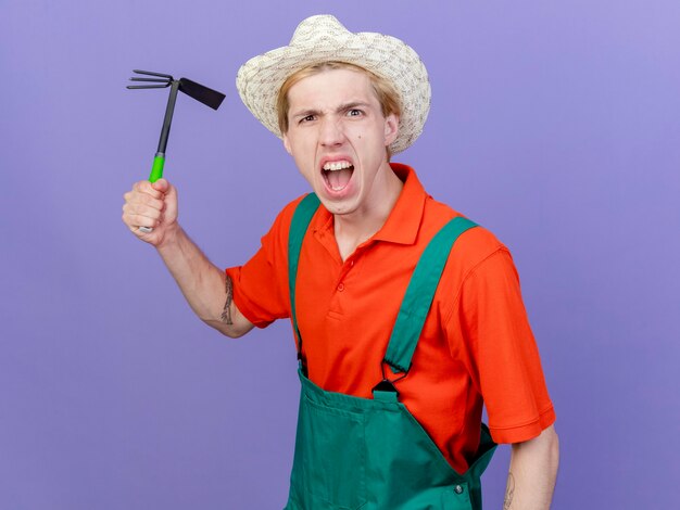 Joven jardinero hombre vestido con mono y sombrero batidor de balanceo con cara enojada