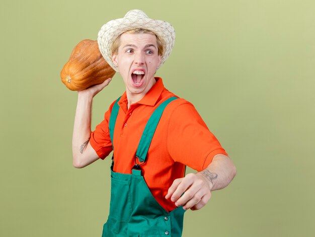 Joven jardinero hombre vestido con mono y sombrero de calabaza balanceándose gritando con expresión agresiva de pie sobre fondo claro