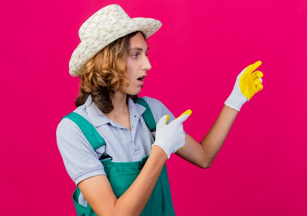 Joven jardinero hombre vestido con mono y sombrero con guantes de goma apuntando