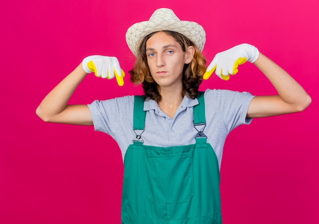 Joven jardinero hombre vestido con mono y sombrero con guantes de goma apuntando