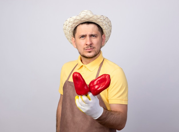 Joven jardinero hombre vestido con mono y sombrero en guantes de trabajo mostrando pimientos rojos mirando confiado