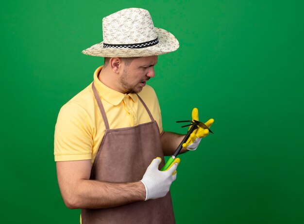 Foto gratuita joven jardinero hombre vestido con mono y sombrero en guantes de trabajo sosteniendo azadón mirándolo con cara seria