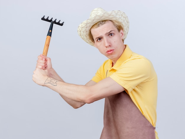 Joven jardinero hombre vestido con mono y sombrero mini rastrillo oscilante está disgustado