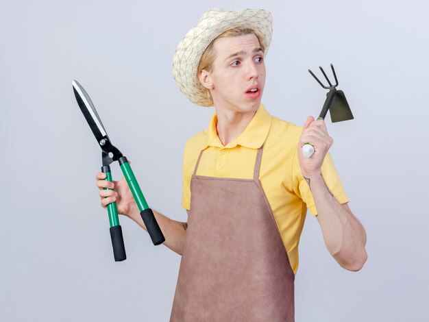 Foto gratuita joven jardinero hombre vestido con mono y sombrero sosteniendo azadón y cortasetos mirando a un lado desconcertado