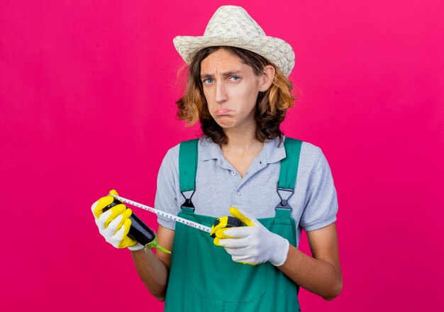 Joven jardinero hombre vestido con mono y sombrero sosteniendo berenjena midiéndolo