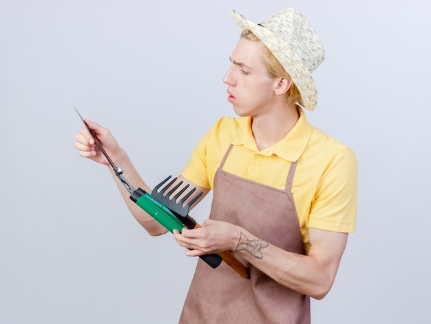 Foto gratuita joven jardinero hombre vestido con mono y sombrero sosteniendo cortasetos y mini rastrillo mirándolos con cara seria