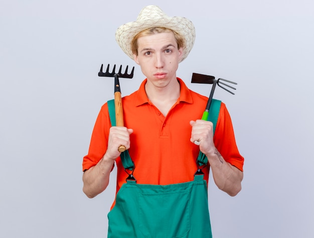 Joven jardinero hombre vestido con mono y sombrero sosteniendo mini rastrillo y azadón mirando a cámara con cara seria de pie sobre fondo blanco.