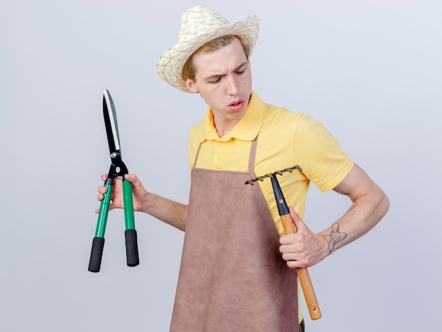 Foto gratuita joven jardinero hombre vestido con mono y sombrero sosteniendo mini rastrillo y cortasetos mirando hacia abajo con cara seria