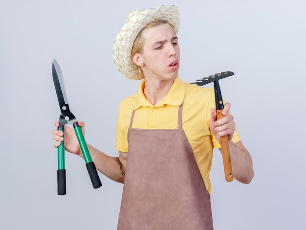 Foto gratuita joven jardinero hombre vestido con mono y sombrero sosteniendo mini rastrillo y cortasetos mirando rastrillo intrigado