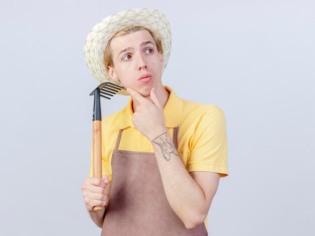 Joven jardinero hombre vestido con mono y sombrero sosteniendo mini rastrillo mirando a un lado desconcertado