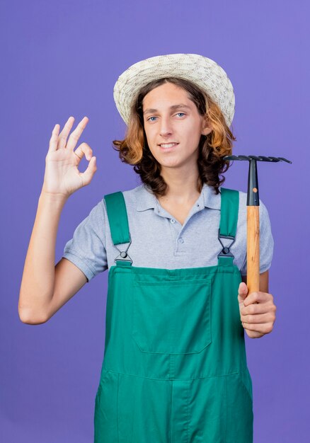 Joven jardinero hombre vestido con mono y sombrero sosteniendo mini rastrillo sonriendo