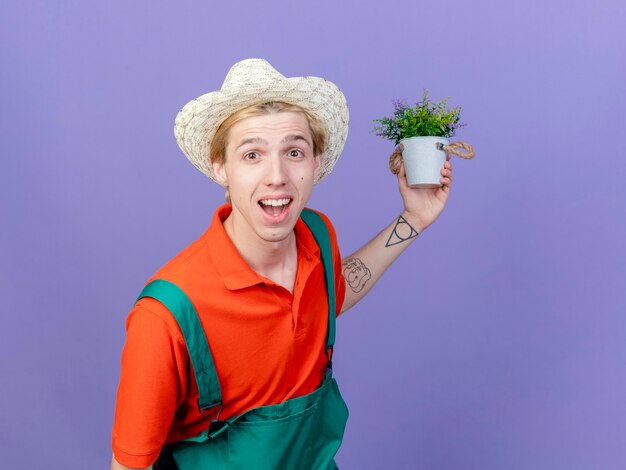 Joven jardinero hombre vestido con mono y sombrero sosteniendo planta en maceta mirando a cámara sonriendo con cara feliz de pie sobre fondo púrpura