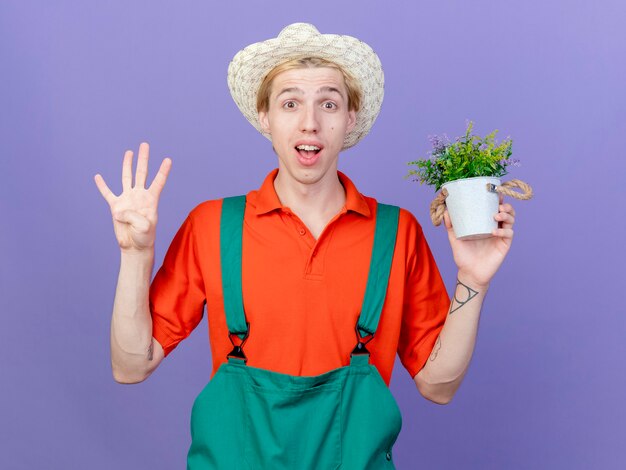 Joven jardinero hombre vestido con mono y sombrero sosteniendo planta en maceta sonriendo