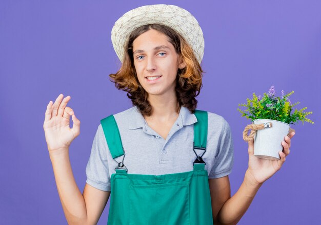 Joven jardinero hombre vestido con mono y sombrero sosteniendo planta en maceta con sonrisa