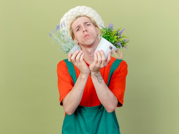 Joven jardinero hombre vestido con mono y sombrero sosteniendo plantas en macetas mirando a la cámara confundido de pie sobre fondo claro