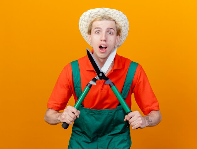 Joven jardinero hombre vestido con mono y sombrero sosteniendo podadoras de setos mirando a la cámara siendo sorprendido y asombrado de pie sobre fondo naranja