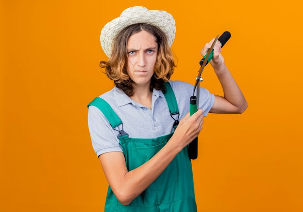 Joven jardinero hombre vestido con mono y sombrero sosteniendo podadoras de setos