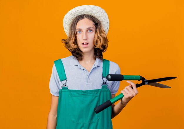 Joven jardinero hombre vestido con mono y sombrero sosteniendo tijeras de podar se confunden