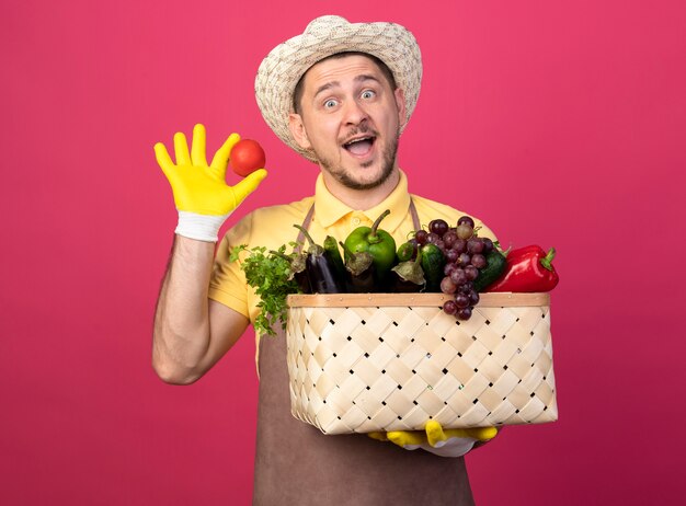 Joven jardinero con mono y sombrero en guantes de trabajo sosteniendo una caja llena de verduras que muestra tomate fresco mirando al frente sonriendo feliz y positivo de pie sobre la pared rosa