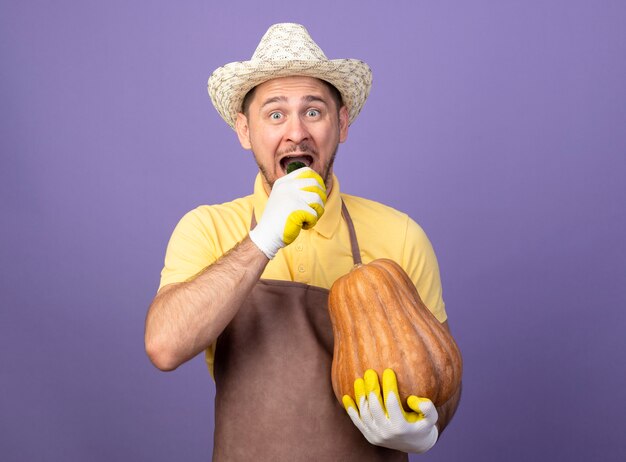 Joven jardinero con mono y sombrero en guantes de trabajo sosteniendo calabaza morder pepino feliz y positivo de pie sobre la pared púrpura