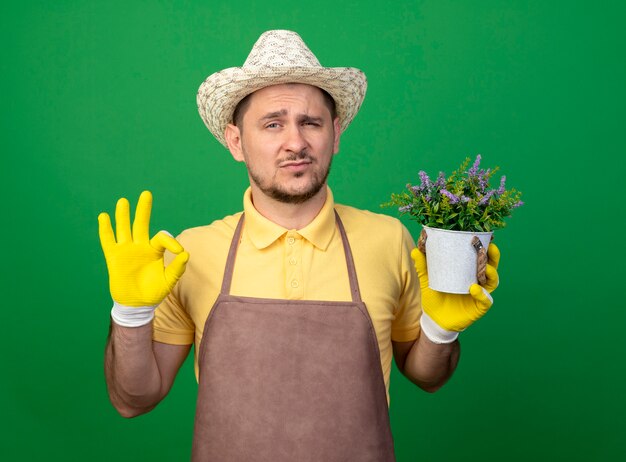 Joven jardinero con mono y sombrero en guantes de trabajo sosteniendo una planta en maceta mirando al frente con expresión de confianza que muestra el signo de ok de pie sobre la pared verde