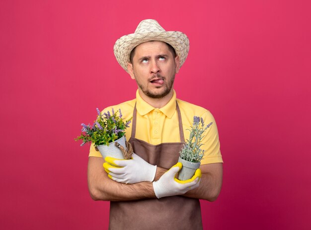 Joven jardinero con mono y sombrero en guantes de trabajo sosteniendo plantas en macetas mirando desconcertado sacando la lengua de pie sobre la pared rosa