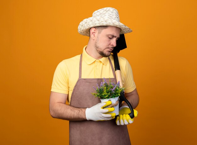 Joven jardinero en mono y sombrero sosteniendo una pala y una planta en maceta mirando hacia abajo con expresión triste de pie sobre la pared naranja