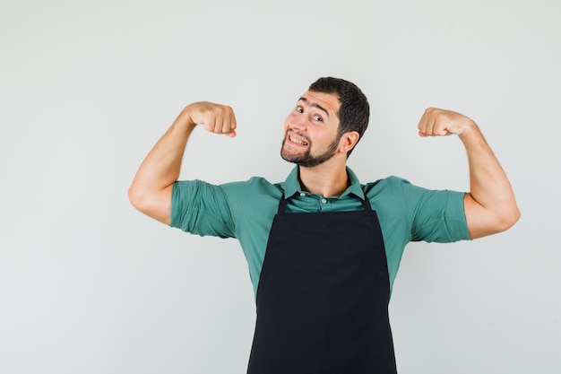 Foto gratuita joven jardinero mostrando los músculos de sus brazos en camiseta, delantal y mirando alegre, vista frontal.