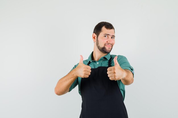 Joven jardinero mostrando el pulgar hacia arriba en camiseta, delantal y mirando contento, vista frontal.