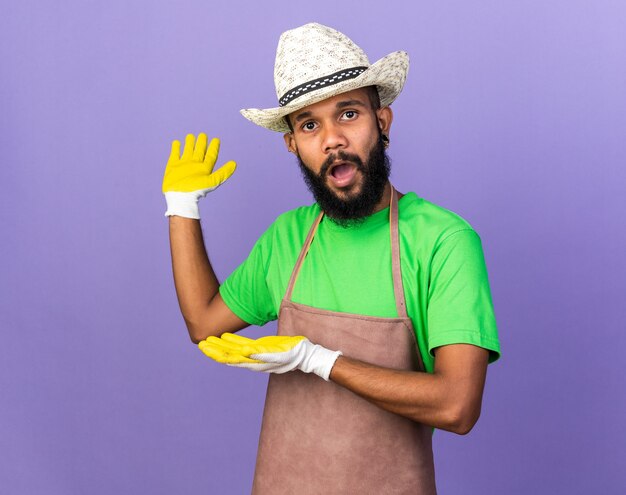 Joven jardinero sorprendido chico afroamericano vistiendo gorro de jardinería con guantes apunta detrás aislado en la pared azul con copia spce