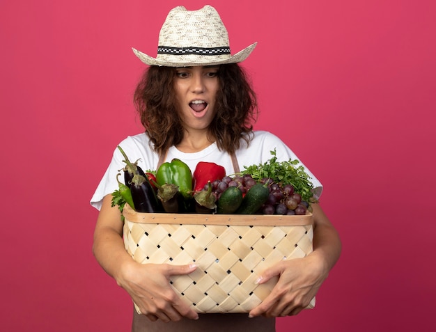 Joven jardinero sorprendido en uniforme con sombrero de jardinería sosteniendo y mirando la canasta de verduras