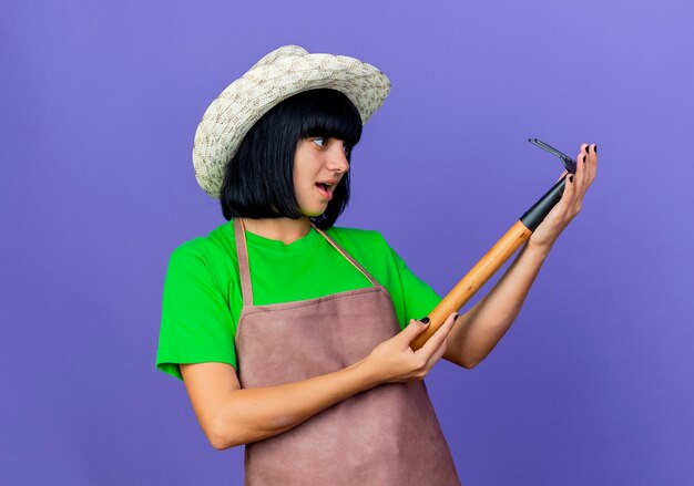 Joven jardinero sorprendido en uniforme con sombrero de jardinería sostiene y mira rastrillo