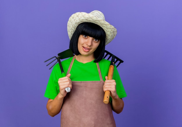 Foto gratuita joven jardinero sorprendido en uniforme con sombrero de jardinería tiene rastrillo y rastrillo azada