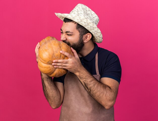 Joven jardinero varón caucásico hambriento vistiendo sombrero de jardinería fingiendo comer calabaza aislado en la pared rosa con espacio de copia