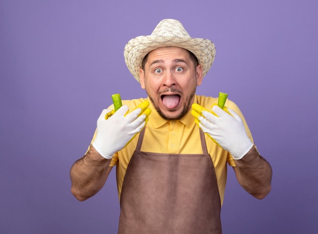 Foto gratuita joven jardinero vestido con mono y sombrero en guantes de trabajo sosteniendo ají verde roto sintiendo que le quema en la boca de pie sobre una pared púrpura