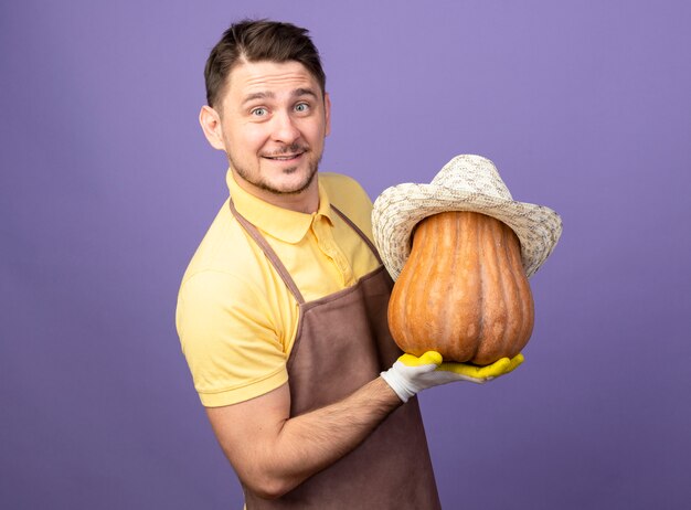 Joven jardinero vistiendo mono en guantes de trabajo sosteniendo calabaza con su sombrero mirando al frente sonriendo con cara feliz de pie sobre la pared púrpura