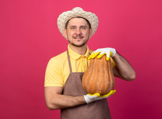 Joven jardinero vistiendo mono y sombrero en guantes de trabajo mostrando calabaza mirando al frente sonriendo confiado de pie sobre la pared rosa