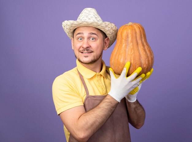 Joven jardinero vistiendo mono y sombrero en guantes de trabajo mostrando calabaza mirando al frente sonriendo feliz y positivo de pie sobre la pared púrpura