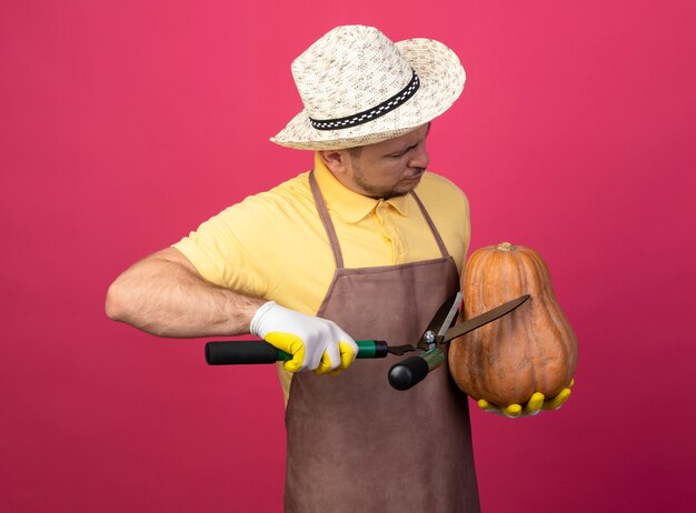 Joven jardinero vistiendo mono y sombrero en guantes de trabajo sosteniendo la calabaza va a cortarla con tijeras de podar de pie sobre una pared rosa