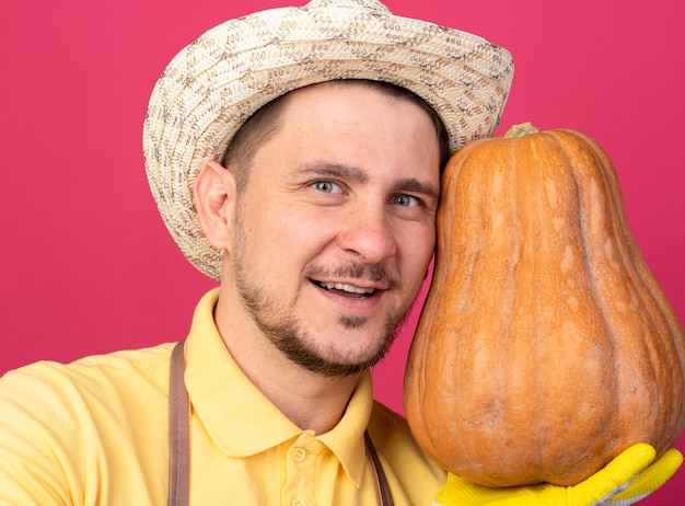 Foto gratuita joven jardinero vistiendo mono y sombrero en guantes de trabajo sosteniendo calabaza mirando al frente sonriendo con cara feliz de pie sobre la pared rosa