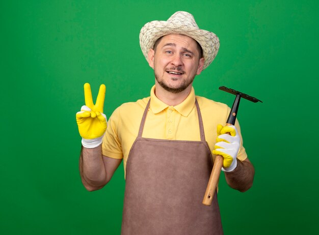 Joven jardinero vistiendo mono y sombrero en guantes de trabajo sosteniendo mini rastrillo mirando al frente v-sign sonriendo de pie sobre la pared verde