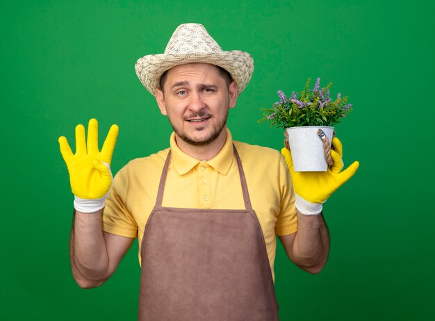 Joven jardinero vistiendo mono y sombrero en guantes de trabajo sosteniendo una planta en maceta mirando al frente mostrando el número cuatro parado sobre la pared verde