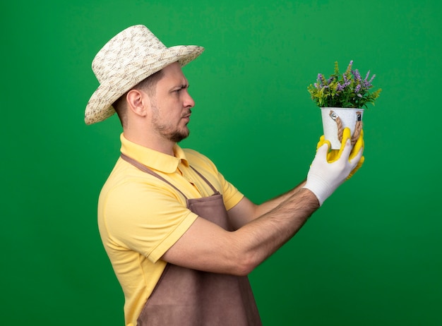 Joven jardinero vistiendo mono y sombrero en guantes de trabajo sosteniendo la planta en maceta mirándola con cara seria de pie sobre la pared verde